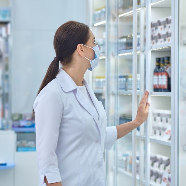 Dark-haired Caucasian female pharmacist in a face mask standing in front of the drugstore display cabinet